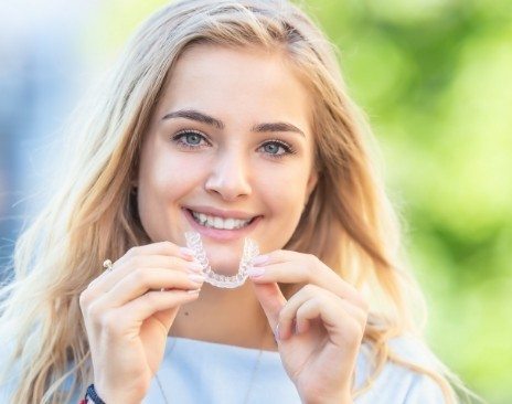 Smiling blonde woman holding a clear aligner near her mouth